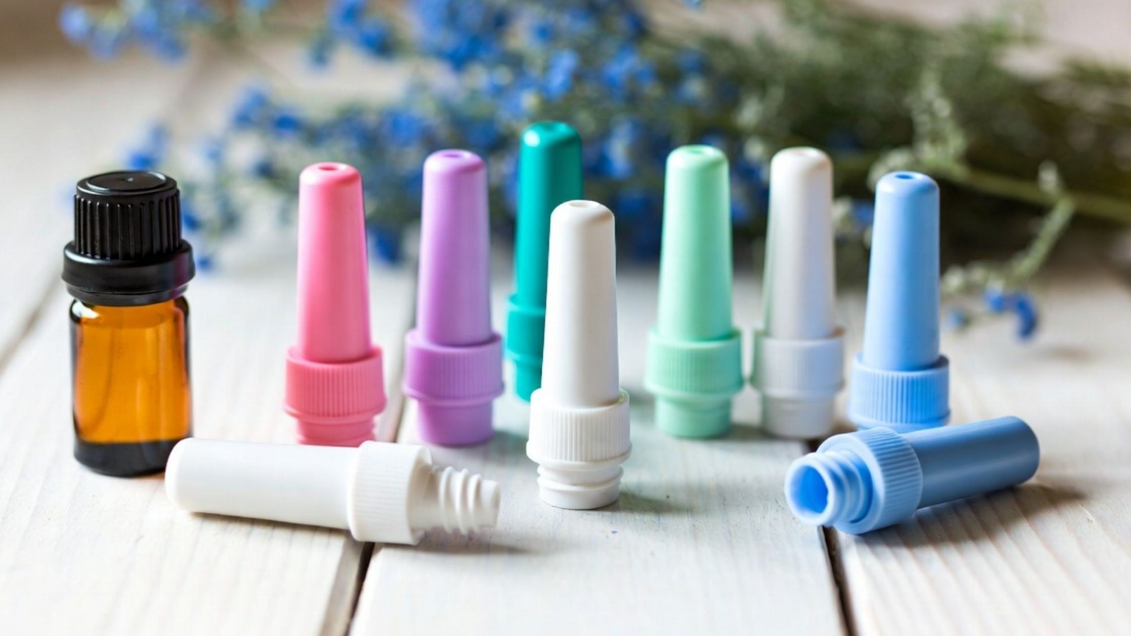 Amber essential oil bottle with colorful nasal inhalers in pink, purple, teal, white and blue arranged on light wooden surface with blurred lavender background for aromatherapy